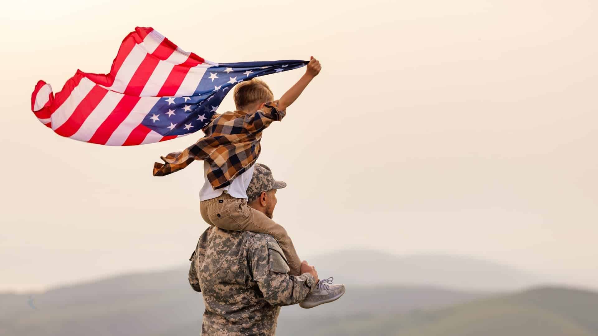 Military personnel walking with child on his shoulders with the child raising an American flag
